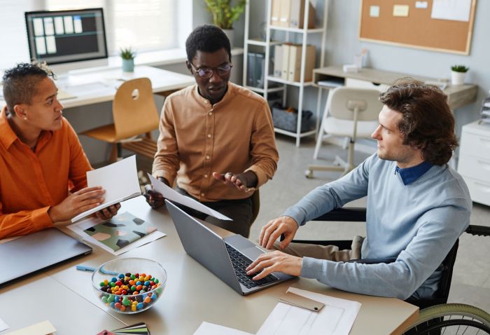 Drei Menschen sitzen an einem Tisch. Ein Mann in einem Rollstuhl an der rechten Bildseite tippt in einen Laptop.